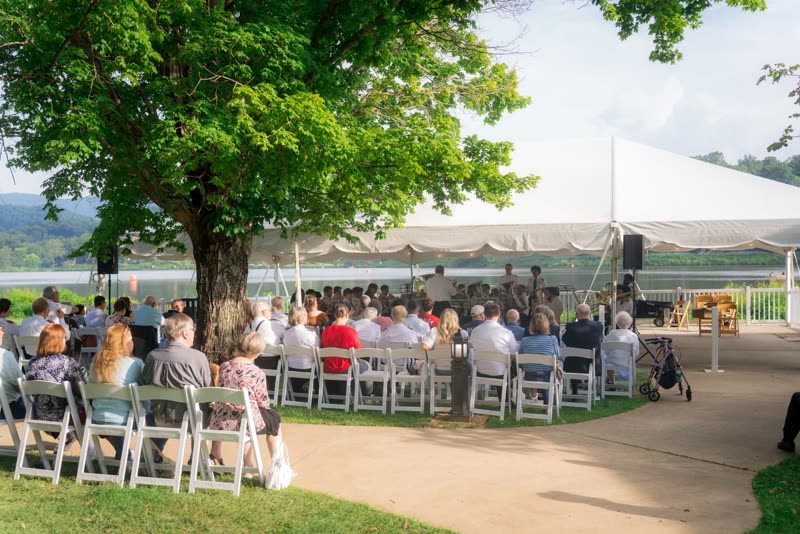 An audience on white folding chairs under trees beside Lake Junaluska, facing a brass band playing under a white marquee