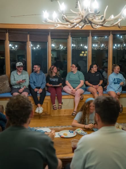 Young adults gathered on a bench and around a table in a lodge lit by an antler chandelier