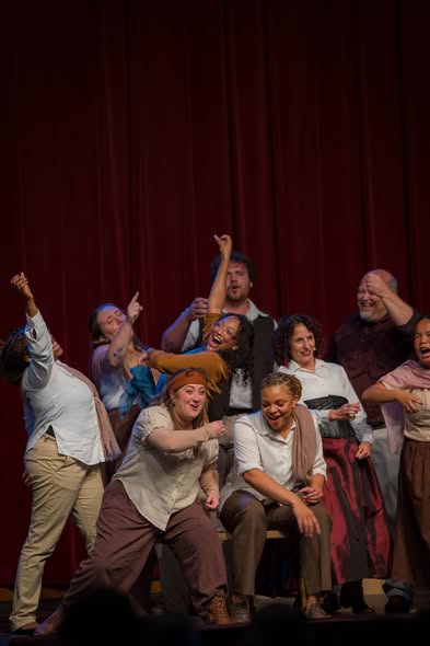 Cast members in period costume performing on stage in front of a red curtain
