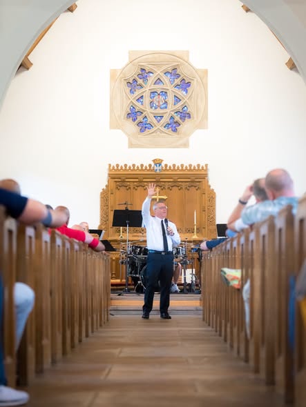A man with his hand raised at the front of a small chapel, seen down the centre aisle