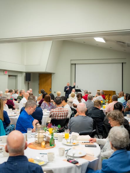 A banquet room full of people at round tables listening to a speaker at the front
