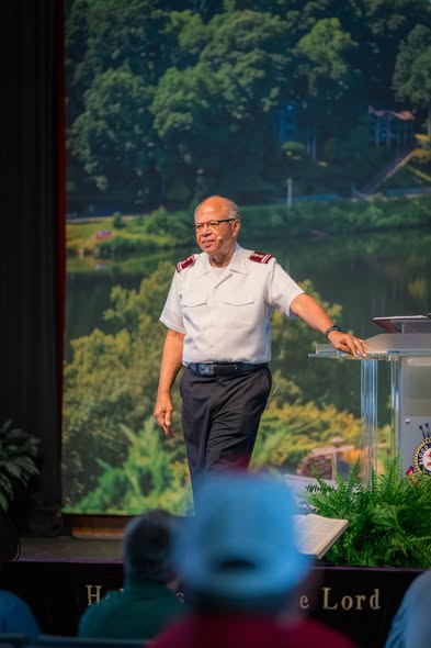 Commissioner Israel Gaither in Salvation Army uniform speaking on stage before a lake backdrop