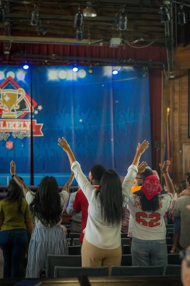 Young people standing with arms raised toward a blue-lit stage beside an "Unlikely" banner