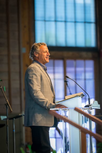 Dr. Thomas McCall speaking from a clear lectern in a timber-framed auditorium