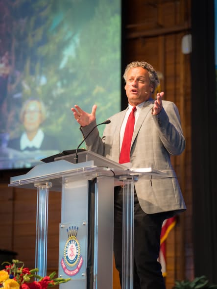 Dr. Thomas McCall gesturing mid-sentence at the lectern with his image on the screen behind