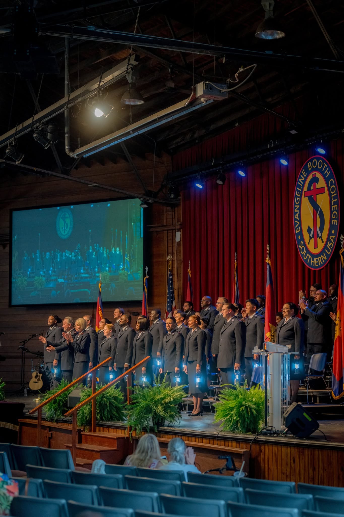 Cadets in uniform standing on stage beneath the Evangeline Booth College crest, one of them holding an orange life ring, with the audience in the foreground
