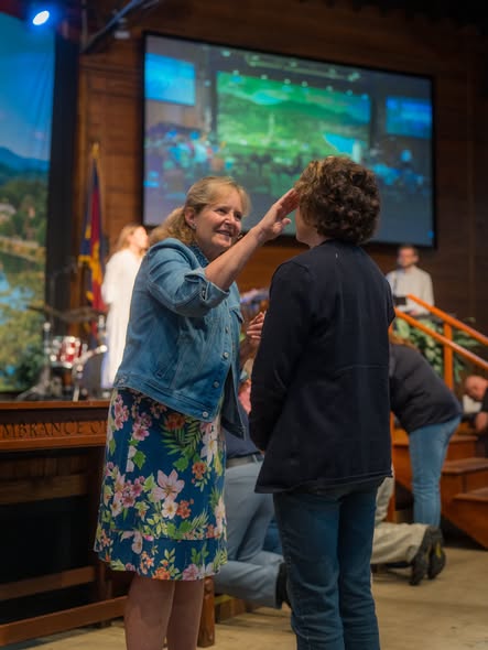 A woman in a denim jacket praying with her hand on another woman’s head at the front of the auditorium