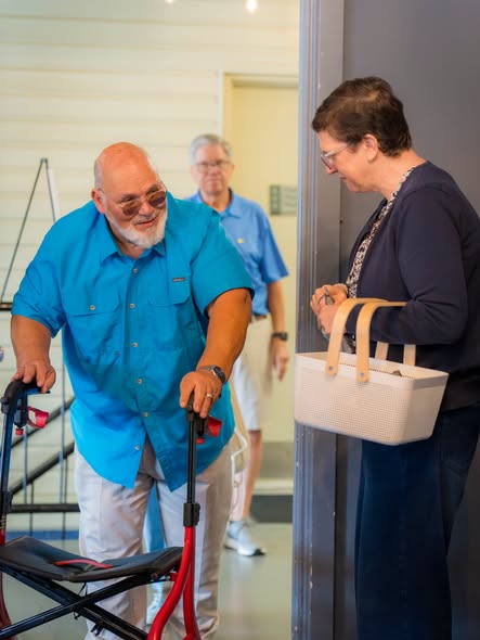 An older man with a walker greeted in a doorway by a woman holding a basket
