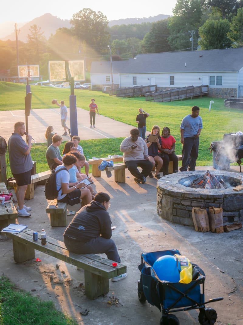 Teens around a stone fire pit and picnic benches at golden hour, basketball court and mountains behind